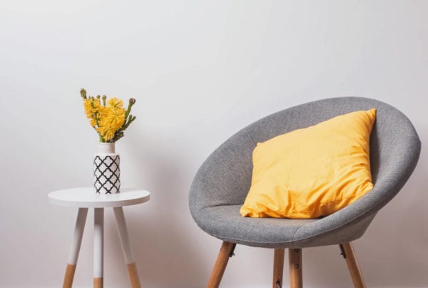 Cozy grey chair with yellow pillow and flowers in the vase standing near the white wall.